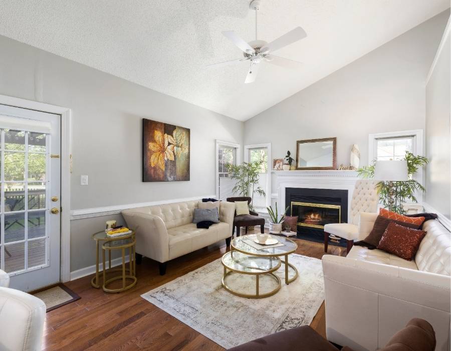 Bright and elegant living room in a Bluffton, SC home featuring white tufted sofas, hardwood floors, glass coffee table, indoor plants, and a cozy fireplace under vaulted ceilings.