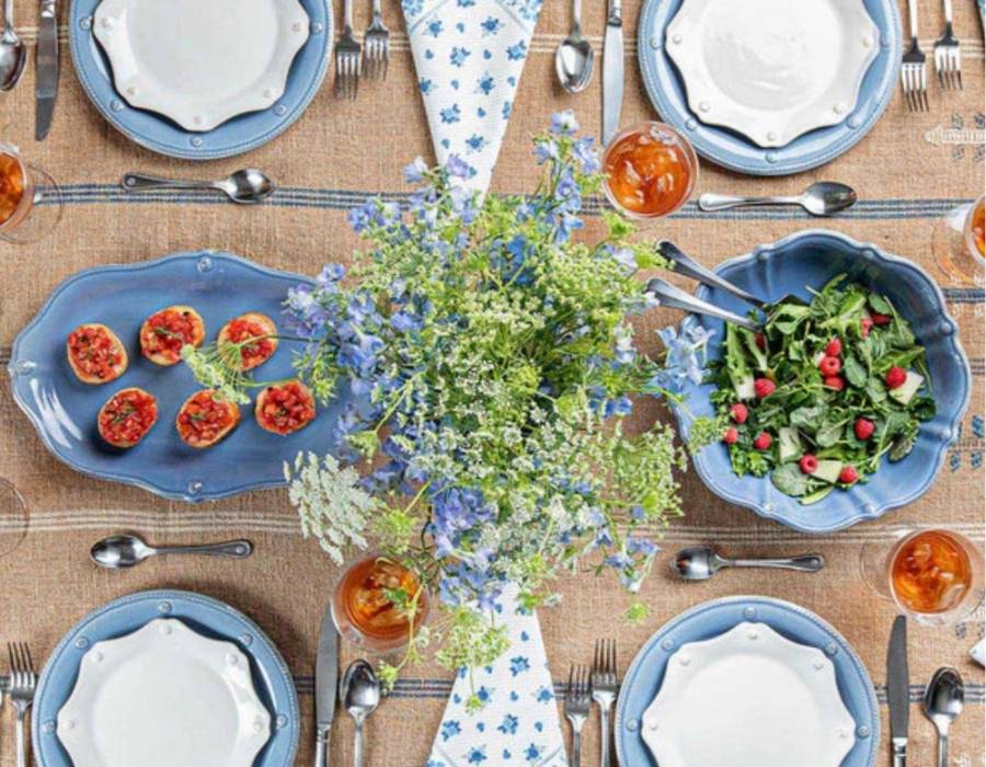 Beautiful farmhouse-style table setting with blue scalloped dinnerware, iced tea, fresh bruschetta, mixed green salad with raspberries, and a vibrant wildflower centerpiece on a rustic burlap tablecloth.