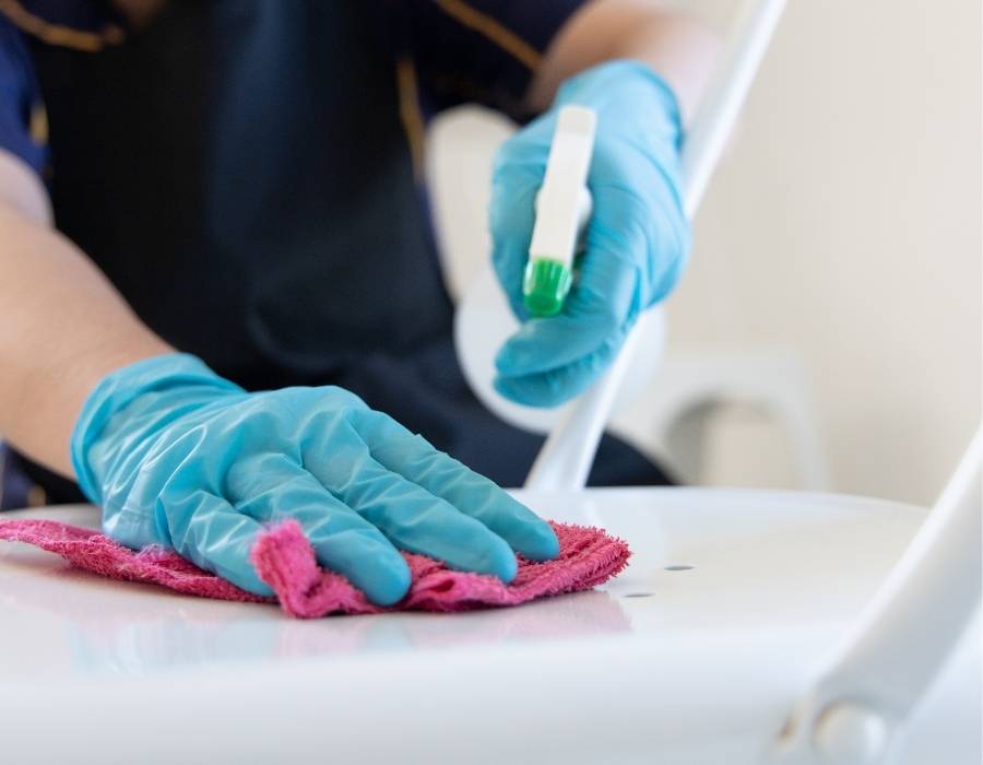 Professional house cleaner wearing blue gloves sanitizing a white chair with disinfectant spray and pink microfiber cloth, representing detailed residential cleaning services.