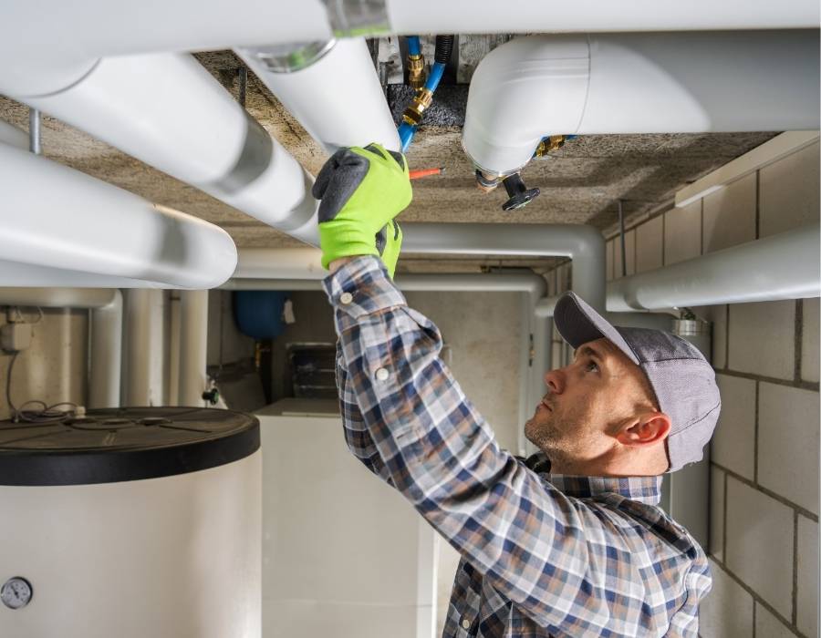 Professional plumber inspecting and servicing insulated piping in a utility room, wearing safety gloves and using tools to perform residential plumbing maintenance.